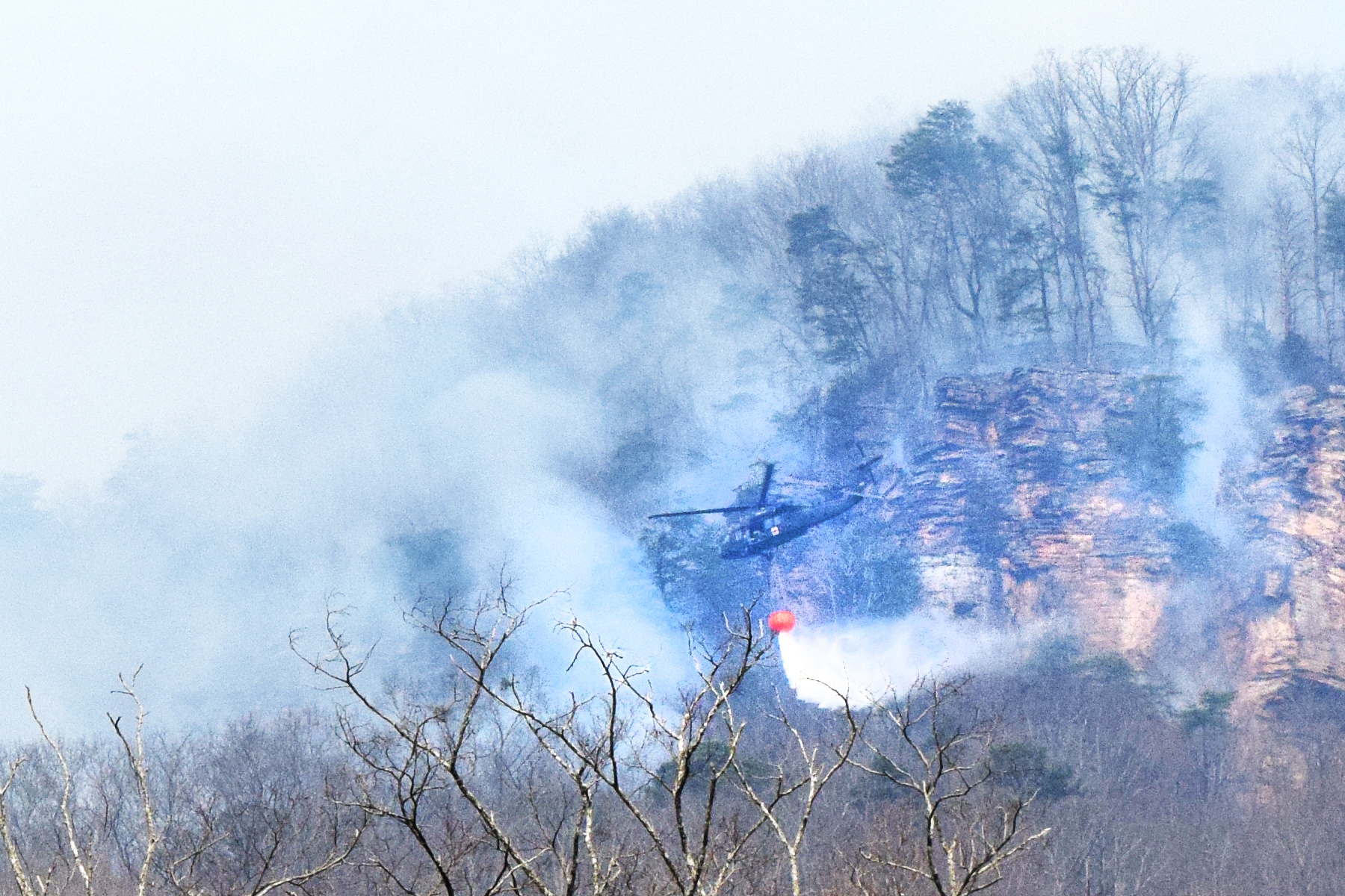 Staff Photo by Ben Benton / A firefighting National Guard helicopter drops a load of water March 13, 2025, on a wildfire in Marion County that has spread across the bluffs north of River Canyon Road near Suck Creek Mountain at the Hamilton County line. The Chattanooga Air Pollution Control Bureau issued a code orange air quality alert related to smoke from area wildfires.