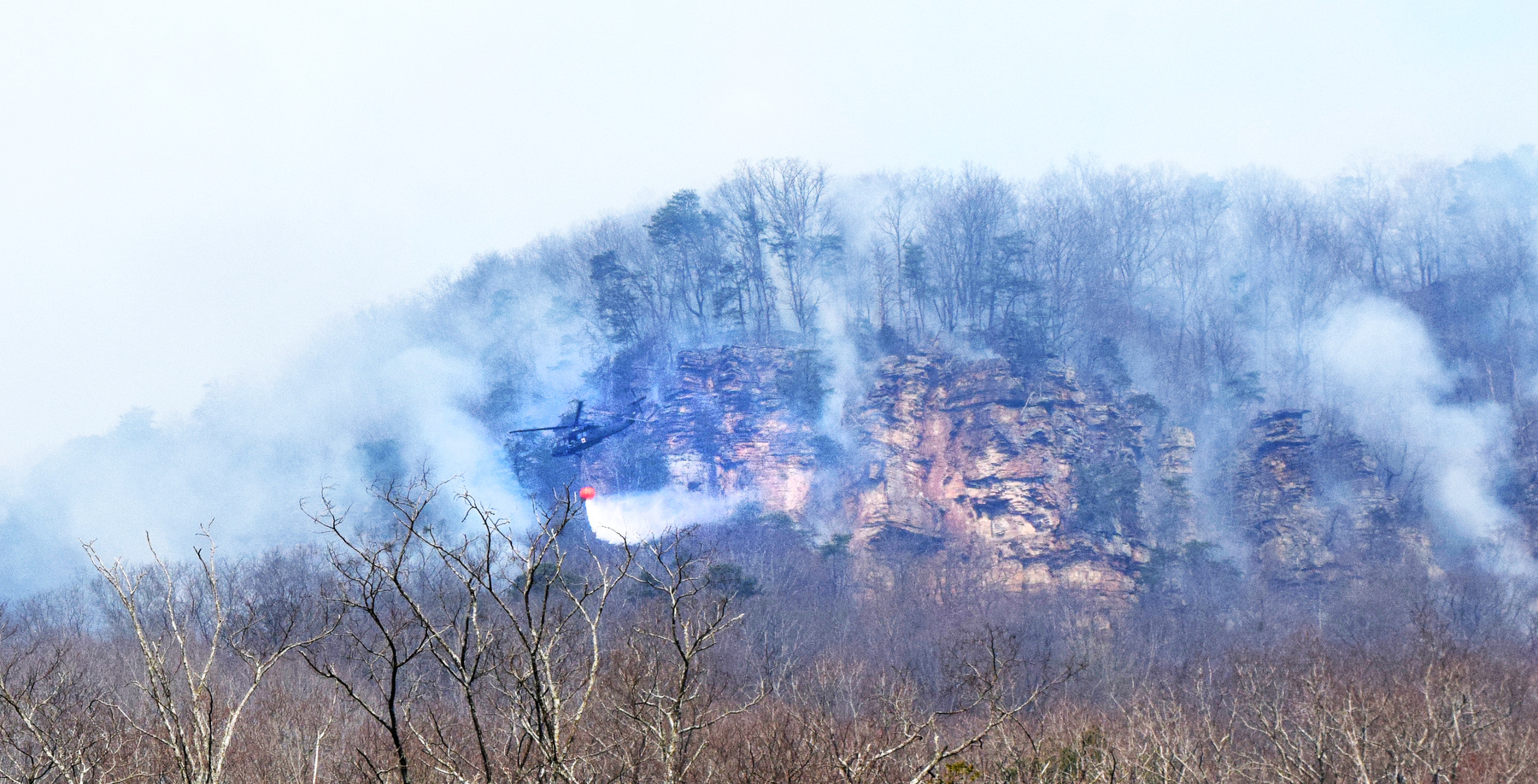 Staff Photo by Ben Benton / A firefighting National Guard helicopter drops a load of water March 13, 2025, on a wildfire in Marion County that has spread across the bluffs north of River Canyon Road near Suck Creek Mountain at the Hamilton County line. The Chattanooga Air Pollution Control Bureau issued a code orange air quality alert related to smoke from area wildfires.