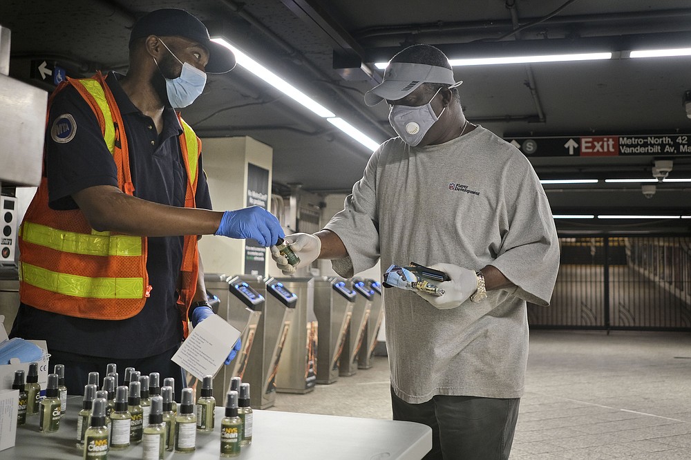 MTA employee Shawn Turner, left, gives away masks and hand sanitizer at the entrance to a subway station in New York, Monday, June 8, 2020. After three months of a coronavirus crisis followed by protests and unrest, New York City is trying to turn a page when a limited range of industries reopen Monday. (AP Photo/Seth Wenig)