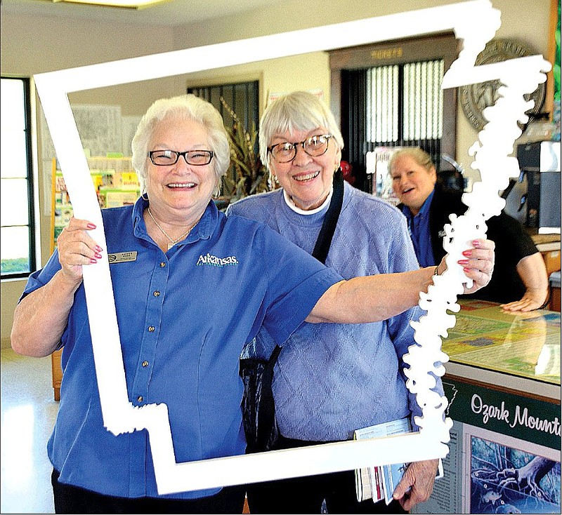 Janelle Jessen/Siloam Sunday Betty Ross, manager of the Arkansas Welcome Center at Siloam Springs, posed with Peggy Wilson of Tahlequah, Okla., with the Arkansas frame the Welcome Center keeps on hand for visitors to take selfies with.