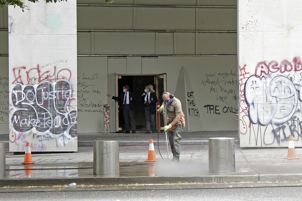 A worker washes graffiti off the sidewalk in front of the Mark O. Hatfield Federal Courthouse in downtown Portland, Ore., on Wednesday, July 8, 2020, as two agents with the U.S. Marshals Service emerge from the boarded-up main entrance to examine the damage. Protesters who have clashed with authorities in Portland, Ore., are facing off not just against city police but a contingent of federal agents who reflect a new priority for the Department of Homeland Security: preventing what President Donald Trump calls "violent mayhem." The agents clad in military-style uniforms include members of an elite Border Patrol tactical unit, and their deployment to protect federal buildings and monuments is a departure for an agency created to focus on threats from abroad. (AP Photo/Gillian Flaccus)