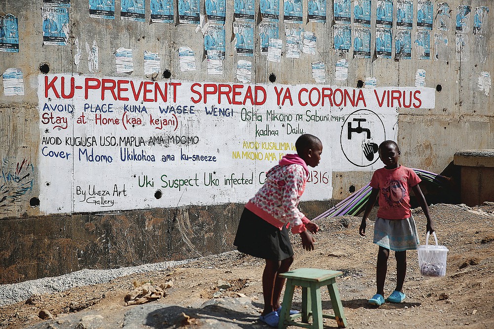 FILE - In this April 2, 2020, file photo, two young girls sell groundnuts in front of an informational mural advising on precautions to avoid catching the new coronavirus, in the Kibera slum, or informal settlement, of Nairobi, Kenya. The options for African students eager to keep studying while schools remain closed because of the coronavirus pandemic seems varied, but the reality for many is that they will fall behind and possibly drop out of school forever, worsening inequality on an already unequal continent. (AP Photo/Brian Inganga, File)