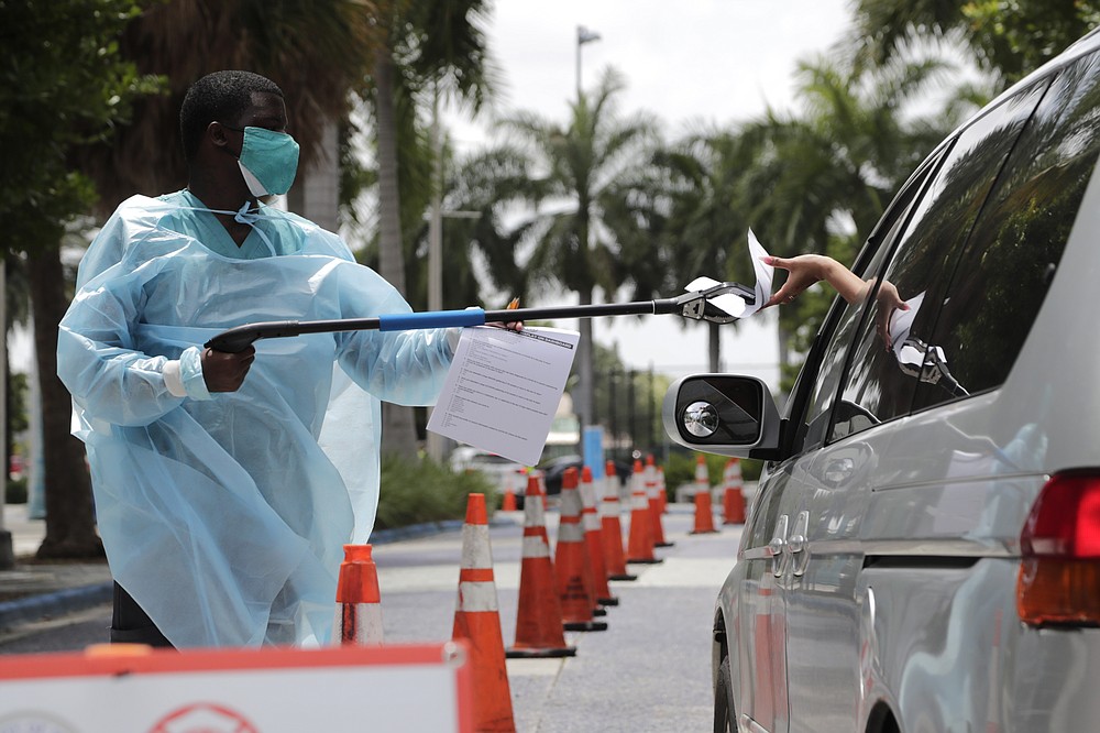 Healthcare worker Dante Hills, left, passes paperwork to a woman in a vehicle at a COVID-19 testing site outside of Marlins Park, Monday, July 27, 2020, in Miami. The Marlins home opener against the Baltimore Orioles on Monday night has been postponed as the Marlins deal with a coronavirus outbreak that stranded them in Philadelphia. (AP Photo/Lynne Sladky)