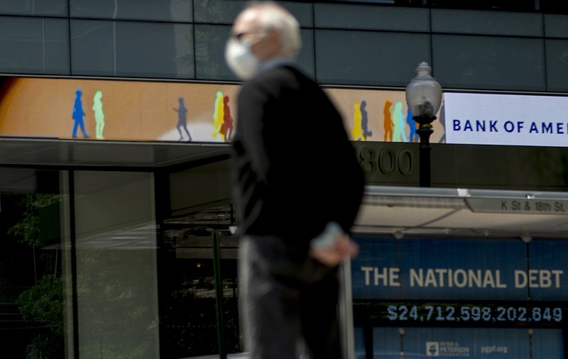 FILE - In this April 29, 2020 file photo, a man wearing a mask to protect against coronavirus, waits to cross the street as a digital sign displays groups of people walking above another sign displaying the size of the national debt along an empty K Street in Washington.  The U.S. budget deficit hit an all-time high of $3 trillion for the first 11 months of this budget year, the Treasury Department said Friday, Sept. 11.  (AP Photo/Andrew Harnik, File)