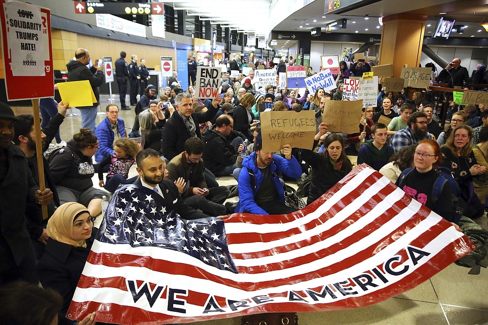 FILE - In this Jan. 28, 2017, file photo, demonstrators sit in the concourse at Seattle-Tacoma International Airport in Seattle, Wash., with a sign that reads "We are America," as more than 1,000 people gather to protest the order signed the day before by President Donald Trump that restricts immigration to the U.S. A federal judge on Thursday, Oct. 1, 2020, temporarily lifted a visa ban on a large number of work permits, undercutting a measure that the Trump administration says will protect American jobs in a pandemic-wracked economy. (Genna Martin/seattlepi.com via AP)