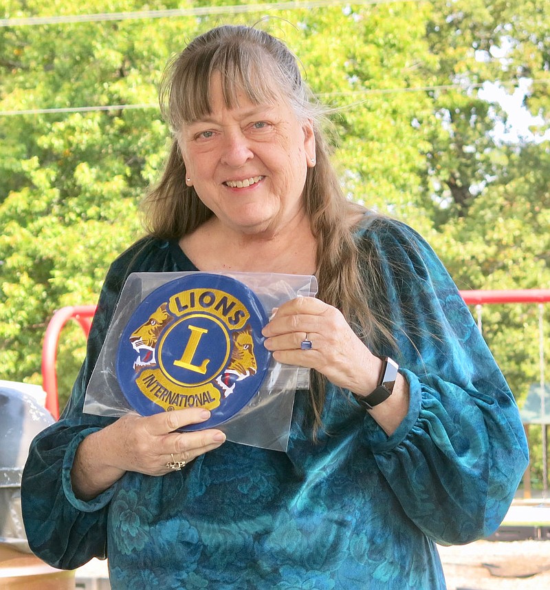 Westside Eagle Observer/SUSAN HOLLAND
Linda Damron, president of the Gravette Lions Club, displays the Lions Club emblem she recently received. The Lions Club has purchased a bench, to be installed on the back deck at the Gravette Public Library, and the patch will be attached to the umbrella which will shade the bench. The bench will provide a convenient place for patrons to read outdoors when the weather is good.