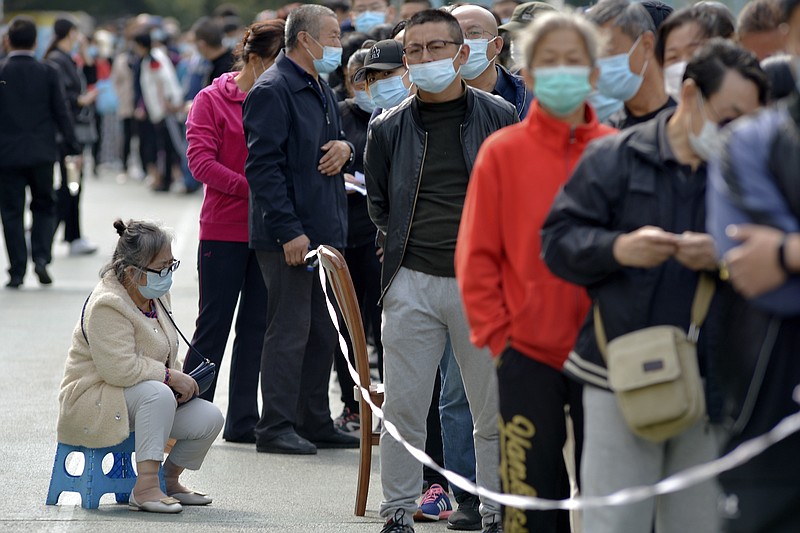 A woman wearing a face mask to help curb the spread of the coronavirus sits on a stool as masked residents line up for the COVID-19 test near the residential area in Qingdao in east China's Shandong province, Monday, Oct. 12, 2020. China's government says all 9 million people in the eastern city of Qingdao will be tested for the coronavirus this week after nine cases linked to a hospital were found. The announcement Monday broke a string of weeks without any locally transmitted infections reported in China. (Chinatopix via AP)