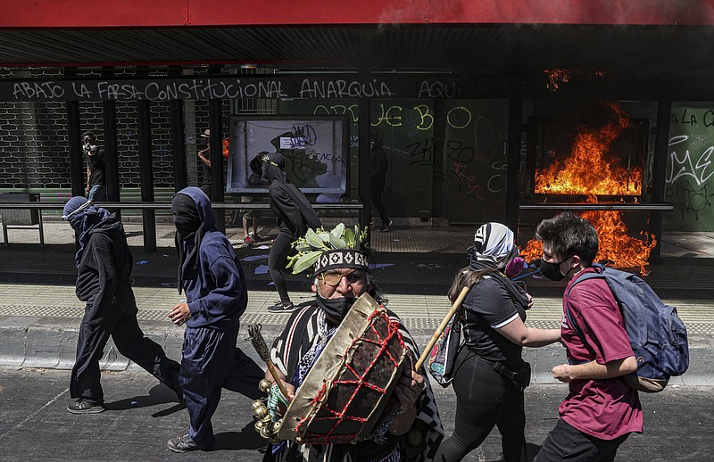 An Indigenous Mapuche man plays a kultrun drum near a bus stop set fire by protesters after police blocked the route of a march against the the discovery of the Americas so it could not pass the presidential palace in Santiago, Chile, Monday, Oct. 12, 2020. The march was organized by Indigenous groups demanding autonomy and the recovery of ancestral land. (AP Photo/Esteban Felix)