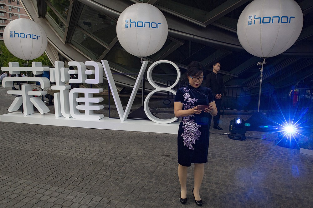 An attendee at a launch event for Huawei's Honor brand mobile phone stands near the brand logo in Beijing, China, Tuesday, May 10, 2016. In an announcement Tuesday, Nov. 17, 2020, Chinese tech giant Huawei says it is selling its budget-price Honor smartphone brand in an effort to rescue the struggling business from damaging U.S. sanctions imposed on its parent company. (AP Photo/Ng Han Guan)