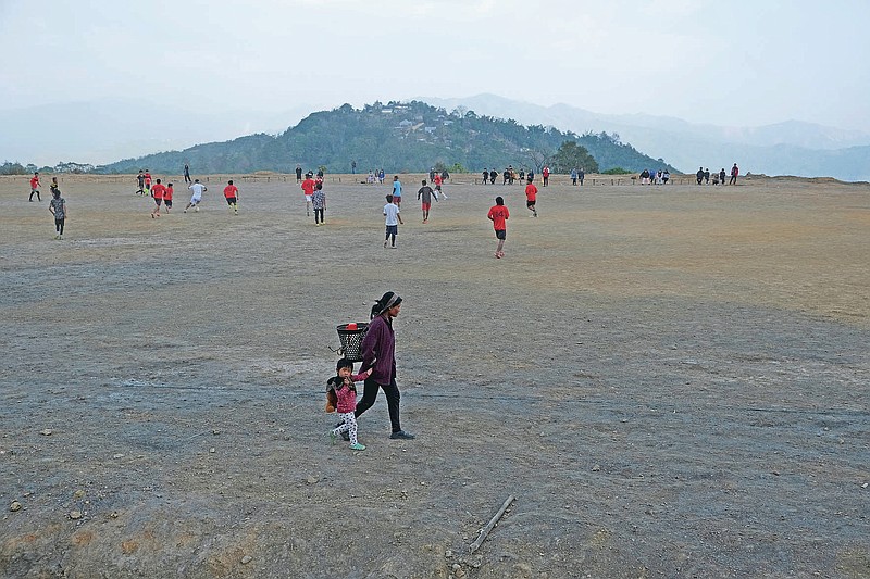 A Tangkhul Naga woman holds the hand of a child and walks past boys engaged in a friendly soccer match, with Nungshang village seen on the mountaintop, in Shangshak village, in the northeastern Indian state of Manipur, Saturday, Jan. 30 2021. Most Naga villages are perched on mountaintops, originally built long ago to spot approaching enemies when the region was little more than a forest. But on nearly every mountaintop, there will be a flat rectangle to serve as a soccer field. (AP Photo/Yirmiyan Arthur)