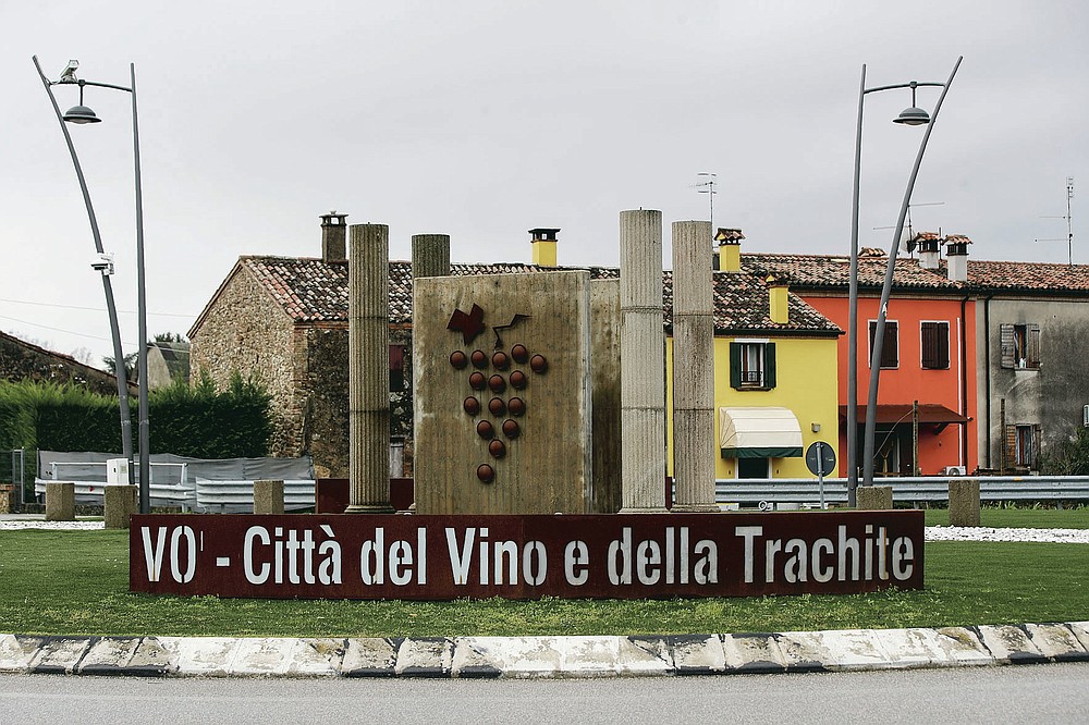 Writing reading in Italian 'Vo - Town of wine and trachyte'' embraces a sculpture on a round-a-bout in town of Vo Euganeo, northern Italy, Wednesday, Feb. 10, 2021. Shockwaves ran through the wine-making village of Vo, west of Venice, as word spread last Feb. 21 that a retired roofer had died of the new coronavirus becoming the first known COVID-19 victim in the West. (AP Photo/Luca Bruno)