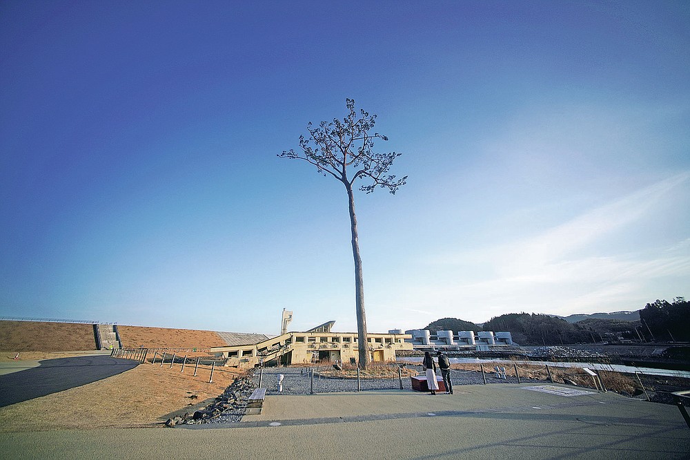 A man and a woman stand near a replica of a lone pine tree that initially survived the 2011 tsunami that flattened the surrounding coastal forest, stands in Rikuzentakata, Iwate Prefecture, northern Japan, Thursday, March 4, 2021. The tree, which eventually died of seawater exposure, was known as the "Miracle Pine," and townspeople treated, reinforced and then preserved it as a memorial and symbol of hope for the region. (AP Photo/Eugene Hoshiko)