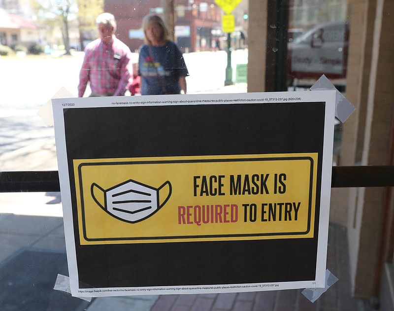 Pedestrians walk along the 600 block of Central Avenue near a sign on a business door requiring face masks for entry Monday. - Photo by Richard Rasmussen of The Sentinel-Record