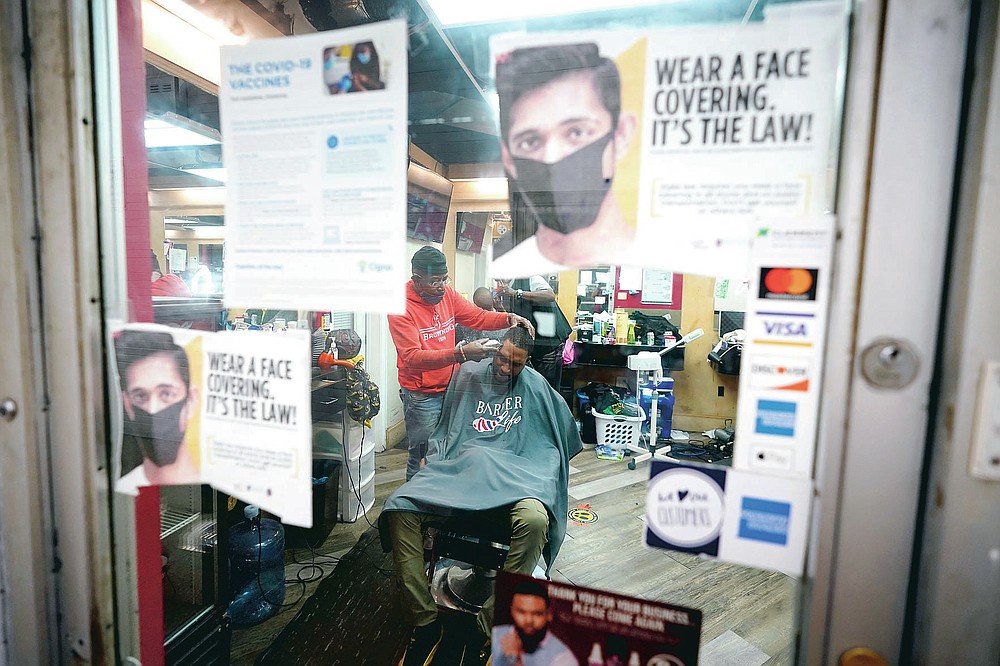 Health literature is posted on a door as Kevin Fitzhugh, center left, cuts the hair of Ikemba Orusakwe, Friday, April 9, 2021, in Hyattsville, Md. Barbers such as Fitzhugh are members of the Health Advocates In Reach & Research (HAIR) program, which helps barbers and hair stylists to get certified to talk to community members about health. During the COVID-19 pandemic, a team of certified barbers have been providing factual information to customers about vaccines, a topic that historically has not been trusted by members of black communities because of the health abuse the race has endured over the years. (AP Photo/Julio Cortez)
