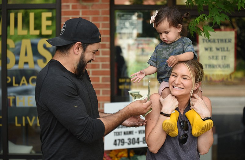 Chase Morman rushes over to catch dollar bills from his daughter Bo, 20 months, on April 28, 2020, as she sits on the shoulders of her mother Kirsten in front of the City Supply store at the downtown square in Fayetteville. The city has formed a steering committee to guide economic recovery from the covid-19 pandemic.
(NWA Democrat-Gazette/David Gottschalk)