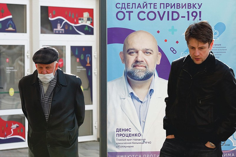 Two men, one of them wearing a face mask, stand near a vaccination point decorated with the poster showing a portrait of Dr. Denis Protsenko and words reading "Get vaccinated against covid-19!!" at VDNKh, The Exhibition of Achievements of National Economy in Moscow, Russia, Friday, April 2, 2021. Moscow is one of the few places in the world where one can get vaccinated against COVID-19 within hours of deciding to do so. Free doses of the domestically developed Sputnik V shot are readily available for anyone 18 or older at more than 200 vaccination points in state and private clinics, shopping malls, food courts, hospitals and even a theater. (AP Photo/Alexander Zemlianichenko)