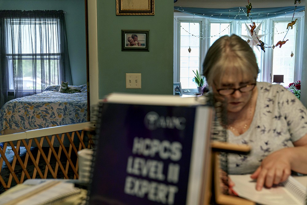 Ellen Booth, 57, studies at her kitchen table to become a certified medical coder as her cat, Juji, sits on a bed behind her in Coventry, R.I., Monday, May 17, 2021. When the restaurant she worked for closed last year, Booth said it gave her "the kick I needed." She started a year-long class to learn to be a medical coder. When her unemployment benefits ran out two months ago, she started drawing on her retirement funds. Booth hopes to pass her upcoming exam and soon hit the job market. (AP Photo/David Goldman)