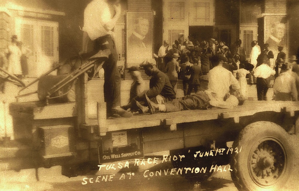 This postcard provided by the Department of Special Collections, McFarlin Library, The University of Tulsa shows a truck parked in front of the Convention Hall, with a man whose condition is unknown, lying on the bed of the truck, and two others sit to either side. A man in civilian attire stands guard over them during the Tulsa Race Massacre June 1, 1921, in Tulsa, Okla. (Department of Special Collections, McFarlin Library, The University of Tulsa via AP)