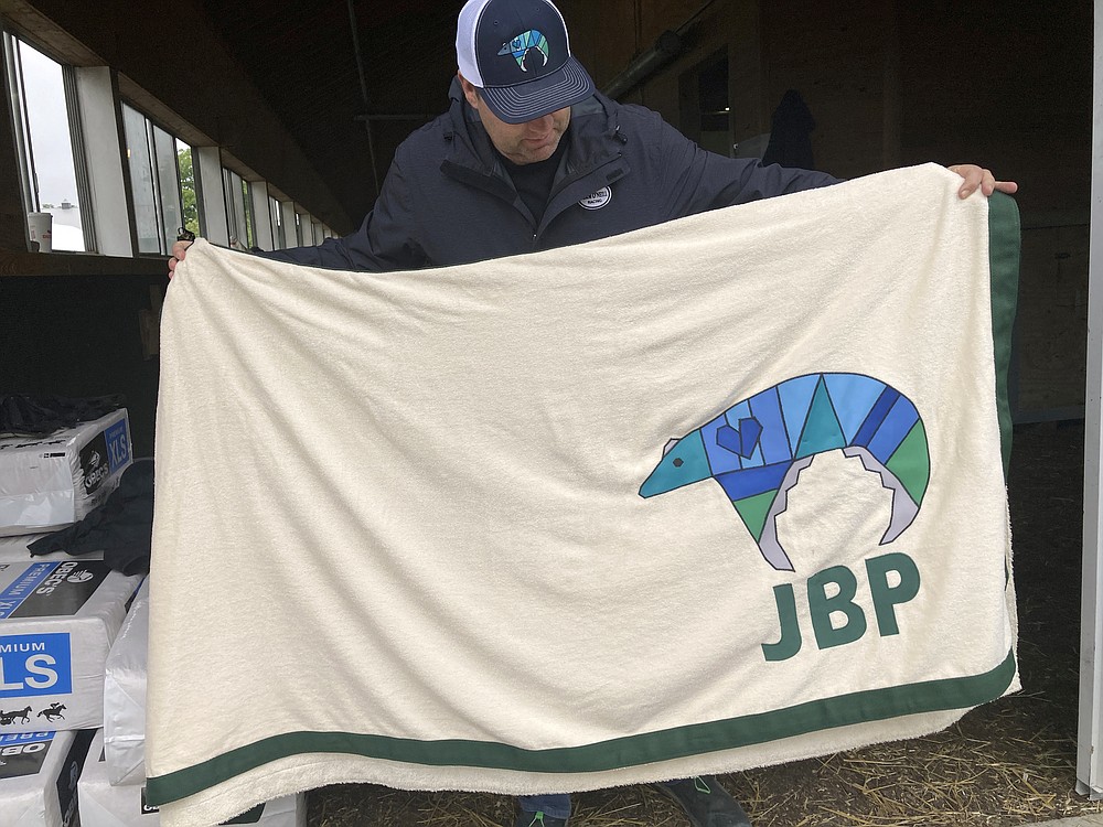 Trainer Doug O’Neill holds up the saddle cloth for Belmont Stakes entrant Hot Rod Charlie at Belmont Park, Thursday, June 3, 2021 , in Elmont, N.Y., featuring the initials of Jake Panus, who was killed in a car crash last year at age 16. O’Neill and some of Hot Rod Charlie’s owners have embraced Panus’ memory. (AP Photo/Stephen Whyno)