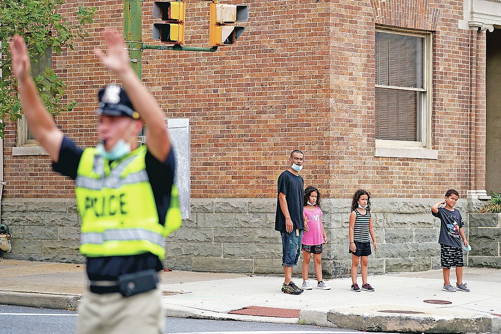 In this Sept. 9, 2020, photo pedestrians look on before crossing an intersection as a cadet in the Baltimore Police Academy directs traffic during an on the field class session in Baltimore. (AP Photo/Julio Cortez)
