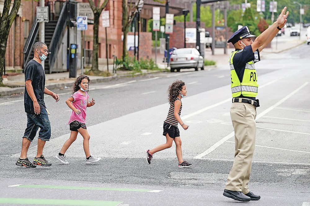 In this Sept. 9, 2020, photo Pedestrians cross an intersection as a cadet in the Baltimore Police Academy directs traffic during an on the field class session in Baltimore. (AP Photo/Julio Cortez)
