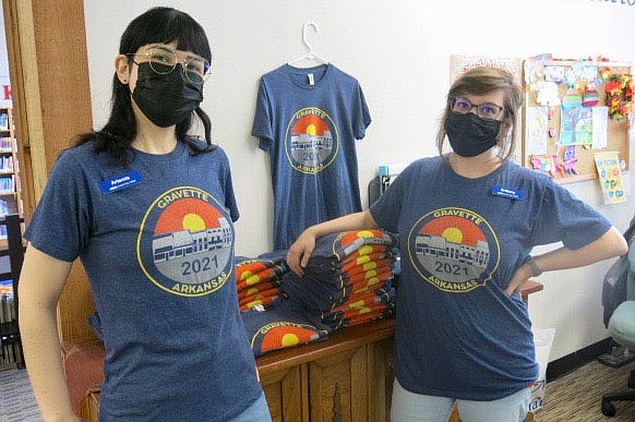 Westside Eagle Observer/SUSAN HOLLAND
Library clerks Artemis Edmisten and Brittany Mangold model 2021 Gravette Day shirts.  The colorful shirts advertising this year's Gravette Day event reflect the 2021 theme "A New Day," with a bright sun rising above Gravette Main Street buildings. The shirts, available in sizes youth large through adult 3X, can be purchased for $12 each at the Gravette Public Library and at City Hall.