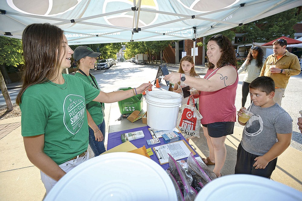 Taylor Gladwin (from left) and Heather Ellzey, both environmental educators for the city of Fayetteville, hand over a bin and food waste collection information on Saturday, July 24, 2021, to Liz Greene of Fayetteville and her children Abram Greene, 6 and Amelia Greene, 11, at the Fayetteville Farmers Market.  The city is encouraging residents to throw away food waste to turn into garbage.  Residents can dump food waste between five locations in the city or on mobile trailers at the Farmers Market every second and fourth Saturday.  Visit nwaonline.com/210725D Daily / for today's photo gallery.  (NWA Democrat-Gazette / Andy Shupe)