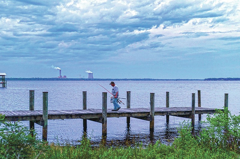 A fisherman walks along a dock on the St. Johns River as a coal-fired power plant stands in the background, in Palatka, Fla., Wednesday, April 14, 2021. After months in a prison cell, Warren Williams longed to fish the St. Johns again. He looked forward to spending days outdoors in his landscaping job, and to writing poems and music in his free time. (AP Photo/David Goldman)