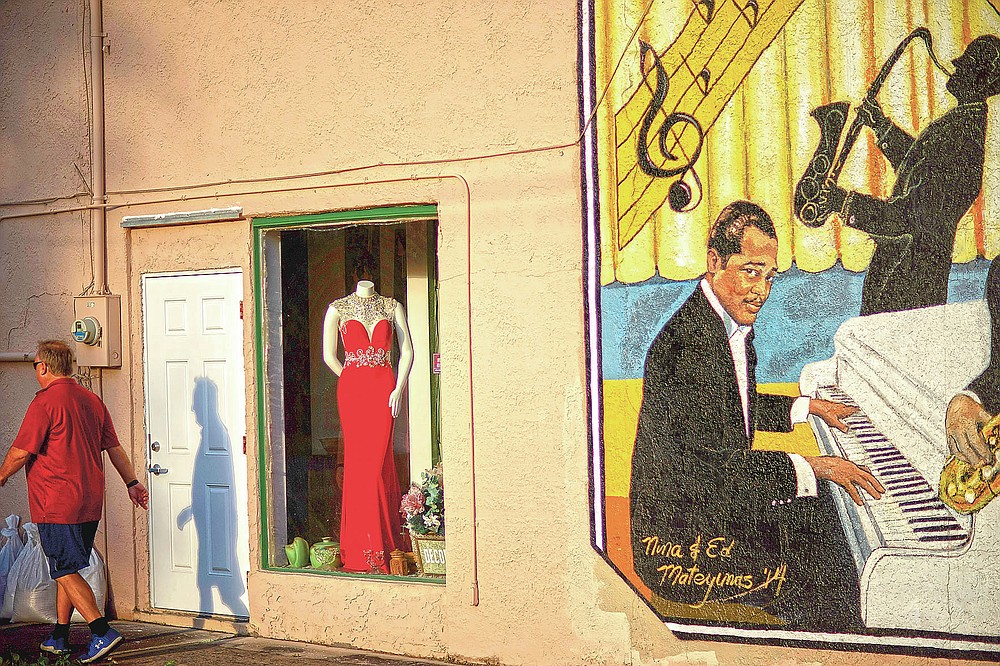 An evening gown is displayed in a store window next to a mural titled "Harlem Nights in Palatka," featuring musicians from the town that formed a 1920s jazz band, as a pedestrian walks by in downtown Palatka, Fla., on Tuesday, April 13, 2021. (AP Photo/David Goldman)