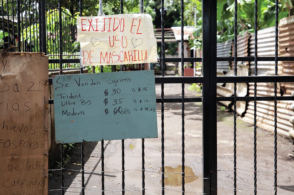 A sign requiring the use of masks during the COVID-19 pandemic hangs over signs advertising cigarettes, eggs, matches, milk and coffee for sale on a gate at the entrance to a residential area in Managua, Nicaragua, Thursday, Sept. 9, 2021. (AP Photo/Miguel Andrés)