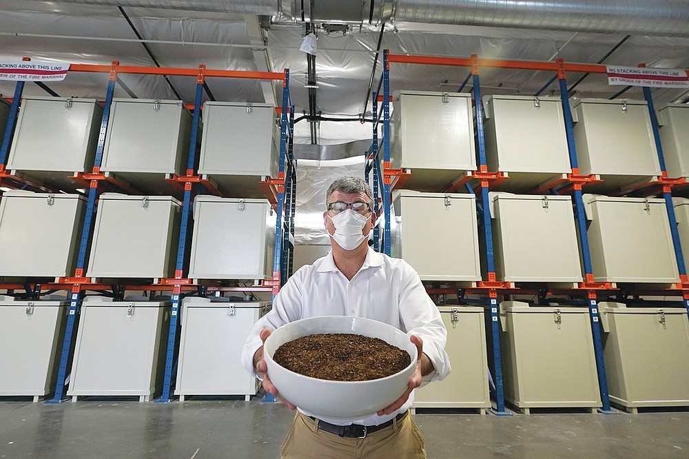Micah Truman, CEO of Return Home, a company that composts human remains into soil, poses for a photo, Wednesday, Sept. 15, 2021, in Auburn, Wash., south of Seattle. Truman is holding a container of soil made with animal remains that is used to show what the product of their process looks like. Behind him are the metal vessels where bodies are kept during the first month of the two-month composing process. Earlier in September, Colorado became the second state after Washington to allow human body composting, and Oregon will allow the practice beginning next July. (AP Photo/Ted S. Warren)