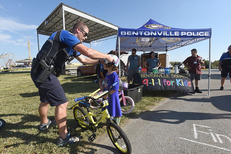 Bentonville police officer Chris Gravley outfits Patience Kwast, 7, with a helmet on Tuesday during the bike rodeo at the Benton County Fair. The department's bike team officers and dispatchers taught bicycle safety and helped kids navigate an obstical course with make-believe fair obstructions such as livestock and cow patties. The rodeo featured a show by bicycle stunt performer Robbie Pfunder of jumps, balancing and other tricks. The fair continues today west of Bentonville. Go to nwaonline.com/210929Daily/ to see more photos.
(NWA Democrat-Gazette/Flip Putthoff)