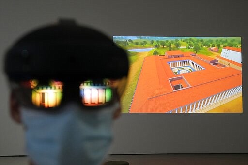 The image of an ancient temple is reflected on a mixed-reality HoloLens headset worn by museum technician Kostas Baskakis as a projection of Ancient Olympia is seen in the background at the Olympic Museum in Athens, Greece, Tuesday, Nov. 9, 2021. The installation is part of a digital restoration project by Microsoft and the Greek Ministry of Culture and Sport at Ancient Olympia, birthplace of the Olympic Games. (AP Photo/Thanassis Stavrakis)