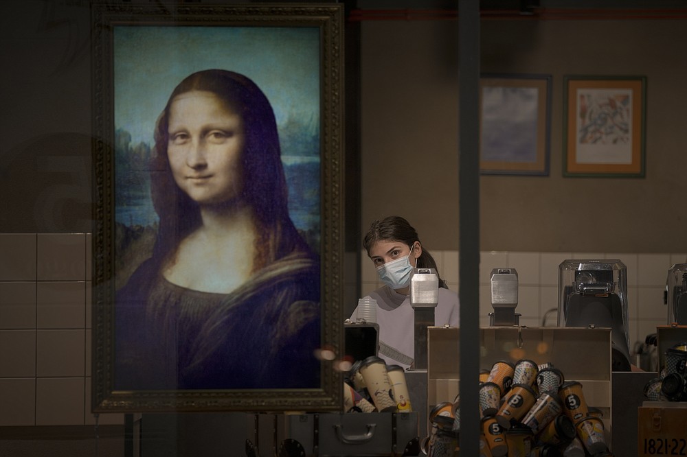 FILE - A display showing an animated depiction of Mona Lisa hangs in a coffee shop window as an employee wearing a mask cleans up shortly after closing hours in Bucharest, Romania, Sunday, Oct. 24, 2021.(AP Photo/Vadim Ghirda, File)