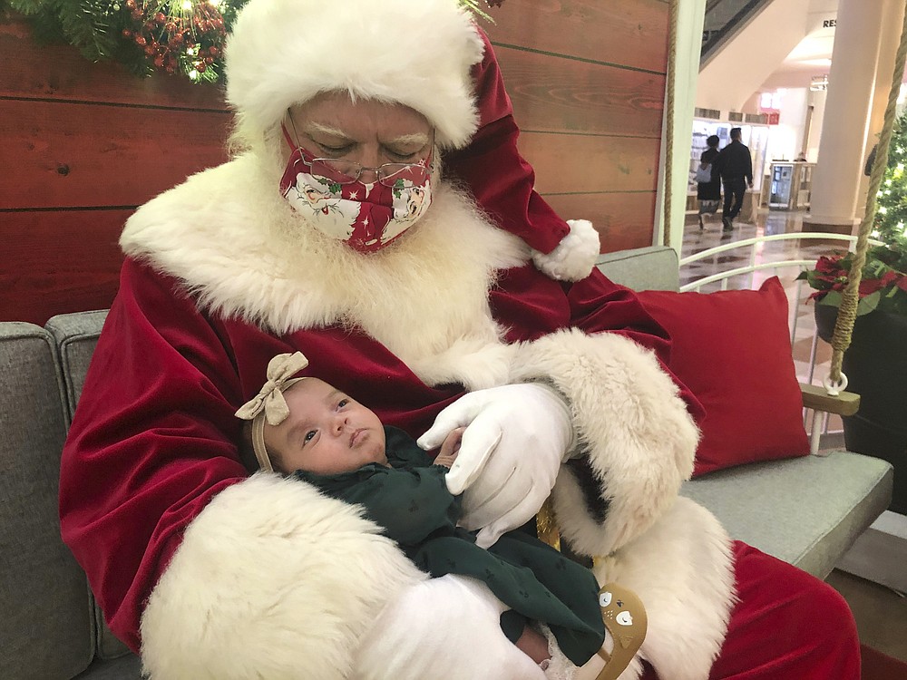 A Santa holds a baby at Stonestown Galleria in San Francisco, on Monday, Nov. 22, 2021. Santa is back this year, but he pleads diligence as he continues to tiptoe through the pandemic. Amid a downturn in available Santas, about 15% fewer in one large database, some are busier than ever. (AP Photo/Haven Daley)