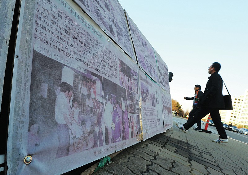 In this April 2, 2019, photo,  people walk past a photo of guards unloading children from a truck at the Brothers Home in Busan, as it is displayed in front of National Assembly in Seoul, South Korea. A notorious South Korean facility that kidnapped, abused and enslaved children, adults and the disabled for a generation was also shipping children overseas for adoption, part of a massive profit-seeking enterprise that thrived by exploiting those trapped within its walls, The Associated Press has found.(AP Photo/Ahn Young-joon)