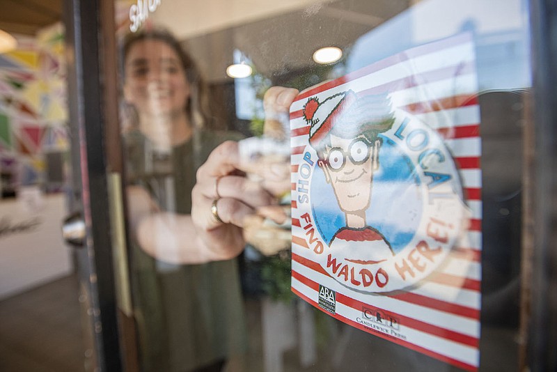 Isla May, owner of Edible Ideaz, affixes a promotional sticker to her store entrance on Friday, July 1, 2022, in downtown Fort Smith. The bakery was one of 17 local businesses and museums that spent the day kicking off the 10th annual Find Waldo Local summer activity, a citywide search in which spotters who locate hidden Waldo figures collect stamps from the participating businesses and exchange them for prizes from host sponsor Bookish: An Indie Shop For Folks Who Read. A drawing for a deluxe set of Waldo books will be held at a grand celebration July 31 at Bookish when the promotion ends. Visit nwaonline.com/220702Daily/ for today's photo gallery.
(NWA Democrat-Gazette/Hank Layton)