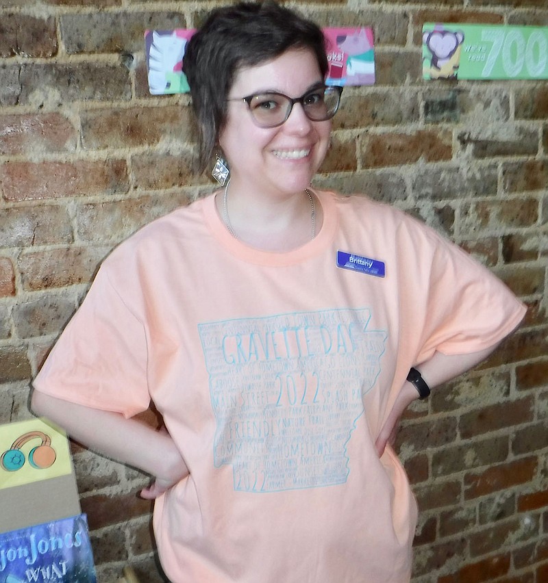 Westside Eagle Observer/SUSAN HOLLAND Library clerk Brittany Mangold wears a big smile as she models the 2022 Gravette Day T-shirt. Shirts are available at City Hall and at the Gravette Public Library while supplies last. Cost is $8 for youth large — adult XL and $10 for adult 2x-3x.