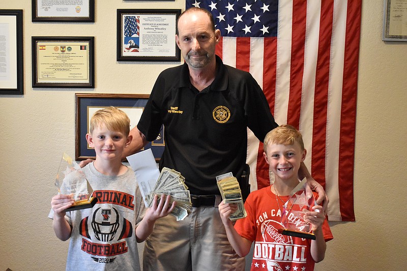 Democrat photo/Garrett Fuller — Ryder Bracht, from left, Moniteau County Sheriff Tony Wheatley and Owen Crane pose together Thursday (Sept. 8, 2022,) as the 8-year-old duo submit their donations for the Shop With A Cop fund at the Moniteau County Sheriff Office. Bracht and Crane, both of Tipton, operated a lemonade stand to raise $1,016. A local employee group matched with a $1,000 donation, resulting in a $2,016 donation to the annual program. Shop With A Cop is an annual event where local law enforcement accompany underprivileged children shopping for Christmas shopping. Donations are accepted by the sheriff office until the day of the event, which is yet to be determined. As a reward for their generosity, the Moniteau County Sheriff Office presented Bracht and Crane each with an acrylic award. They will also get to shop with a cop again this year.