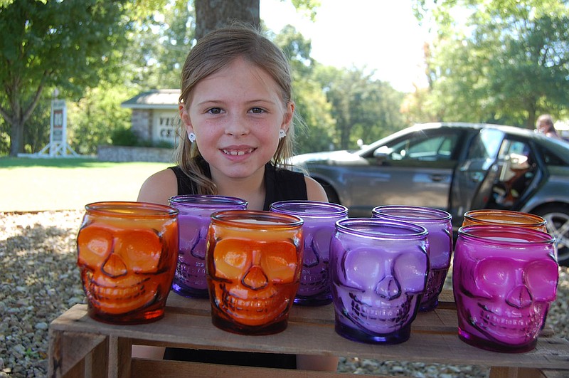 Bennett Horne/The Weekly Vista Addison Greene, 8, smiles from behind a row of Black Sea Skull Candles sold by her mom, Nicole, on Sunday at the Bella Vista Farmers Market.