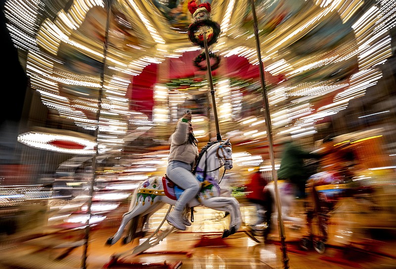FILE - A girls uses her mobile phone as she rides on a horse of a merry-go-round on the Christmas Market in Frankfurt, Germany, Nov. 25, 2022. In cities across Europe, officials are wrestling with a choice this Christmas. Dim lighting plans to send a message of energy conservation and solidarity with citizens squeezed by both higher energy costs and inflation or let the lights blaze in a message of defiance after two years of pandemic-suppressed Christmas seasons, creating a mood that retailers hope loosen holiday purses. (AP Photo/Michael Probst, File)
