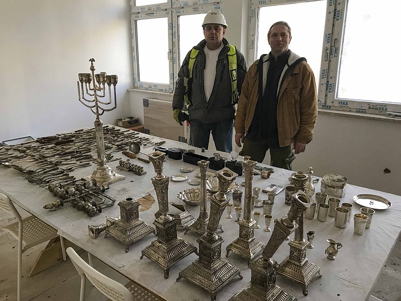 Krzysztof Hejmanowski, left, building inspector of the Warbud construction company and archaeologist Bartlomiej Gwozdz, right, &#xa0;pose for a photograph with objects that were most probably hidden by their Jewish owners during World War II, &#xa0;in Lodz, Poland, Wednesday, Jan. 11, 2023. Lodz city authorities were showing to The Associated Press and commenting about some 400 items - including silver-plated menorahs, hanukkiahs, tableware and daily use items, that were uncovered there last month during house and yard renovation works. (AP Photo/Rafal Niedzielski)