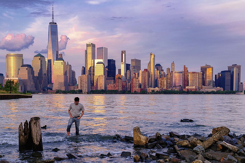 A man checks his footing as he wades through the Morris Canal Outlet on May 31, 2022, in Jersey City, N.J., as the sun sets on the lower Manhattan skyline of New York City. If rising oceans aren't worry enough, add this to the risks New York City faces: The metropolis is sinking under the weight of its skyscrapers, apartment buildings, asphalt and humanity itself — and will eventually become flooded by the Hudson River and Atlantic Ocean. (AP Photo/J. David Ake, File)
