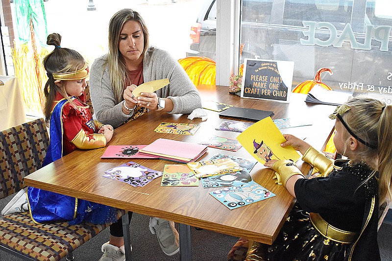 Democrat photo/Garrett Fuller — Paisley Mitchell, right, 4, watches as mother Jessica Mitchell, center, helps younger sister Ellie Mitchell, 2, complete a sticker craft project Friday during the Moniteau County Library's Halloween party.