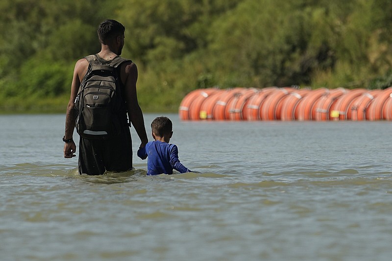 FILE - Migrants walk past large buoys being used as a floating border barrier on the Rio Grande, Aug. 1, 2023, in Eagle Pass, Texas. Texas must move a floating barrier on the Rio Grande that drew backlash from Mexico, a federal appeals court ruled Friday, Dec. 1, 2023, dealing a blow to one of Republican Gov. Greg Abbott's aggressive measures aimed at stopping migrants from entering the U.S. illegally. (AP Photo/Eric Gay, file)