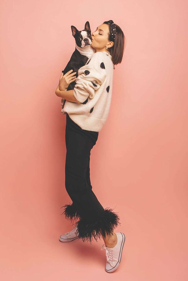 Full length young caucasian woman kissing her black and white dog while standing on pink background. Brunette with bob haircut wears sweater and pants. True friendship, wonderful happy moments