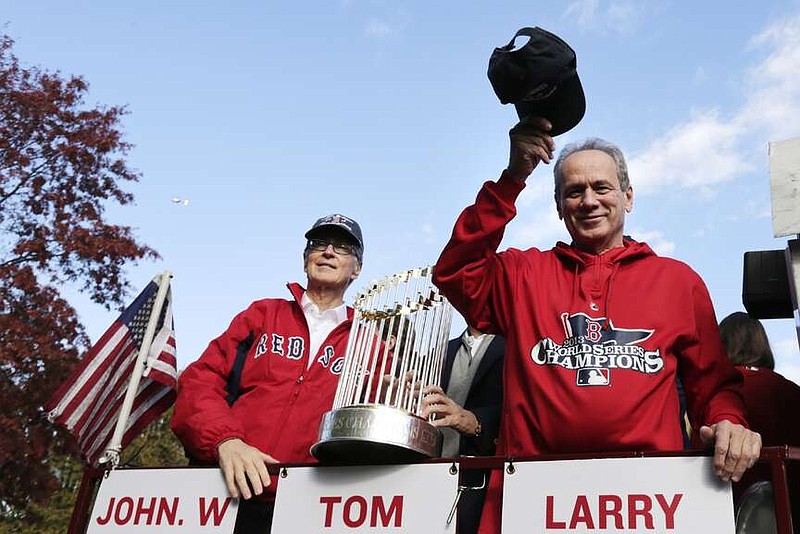 FILE - Boston Red Sox president Larry Lucchino, right, tips his cap to fans as majority owner John Henry holds the 2013 World Series championship trophy during a parade in celebration of the baseball team's win, Saturday, Nov. 2, 2013, in Boston. Larry Lucchino, the force behind baseball's retro ballpark revolution and the transformation of the Boston Red Sox from cursed losers to World Series champions, has died. He was 78. Lucchino had suffered from cancer. The Triple-A Worcester Red Sox, his last project in a career that also included three major league baseball franchises and one in the NFL, confirmed his death on Tuesday, April 2, 2024. (AP Photo/Charles Krupa, File)