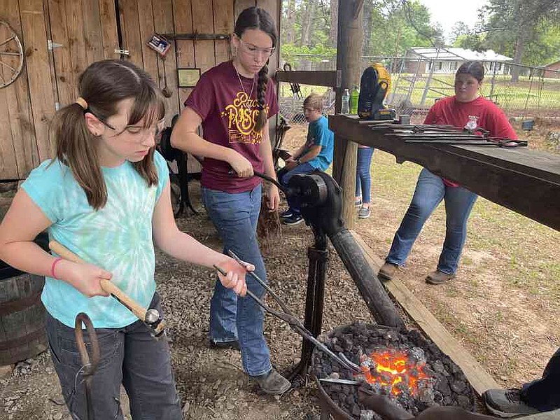 Sadie Montgomery, left, returns her tent stake to the fire to get it ready to shape again, while Kady Jo Strother turns the forge's crank to fan its flames. (Special to The Commercial/University of Arkansas System Division of Agriculture)