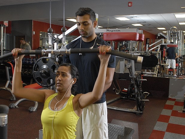 Devraj Patel, standing, assists Deepa Saha as they work with weights at the Workout Anytime fitness center in Hixson.