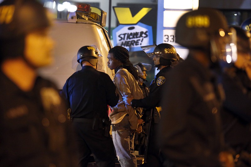 A man is detained by Oakland police officers as other stand guard by Broadway Auto Row where several auto dealerships windows and cars were smashed during one of the May Day protests in Oakland, Calif., on May 1, 2015. (Ray Chavez/The Oakland Tribune via AP)